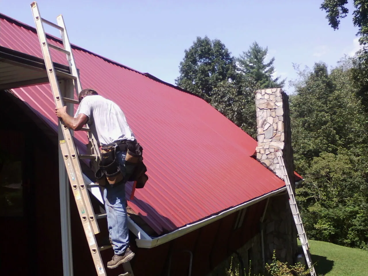 Roofer on a ladder beside a red metal roof and stone chimney — Soffit & Fascia Repair by Roofing Brothers in Atlantic Highlands